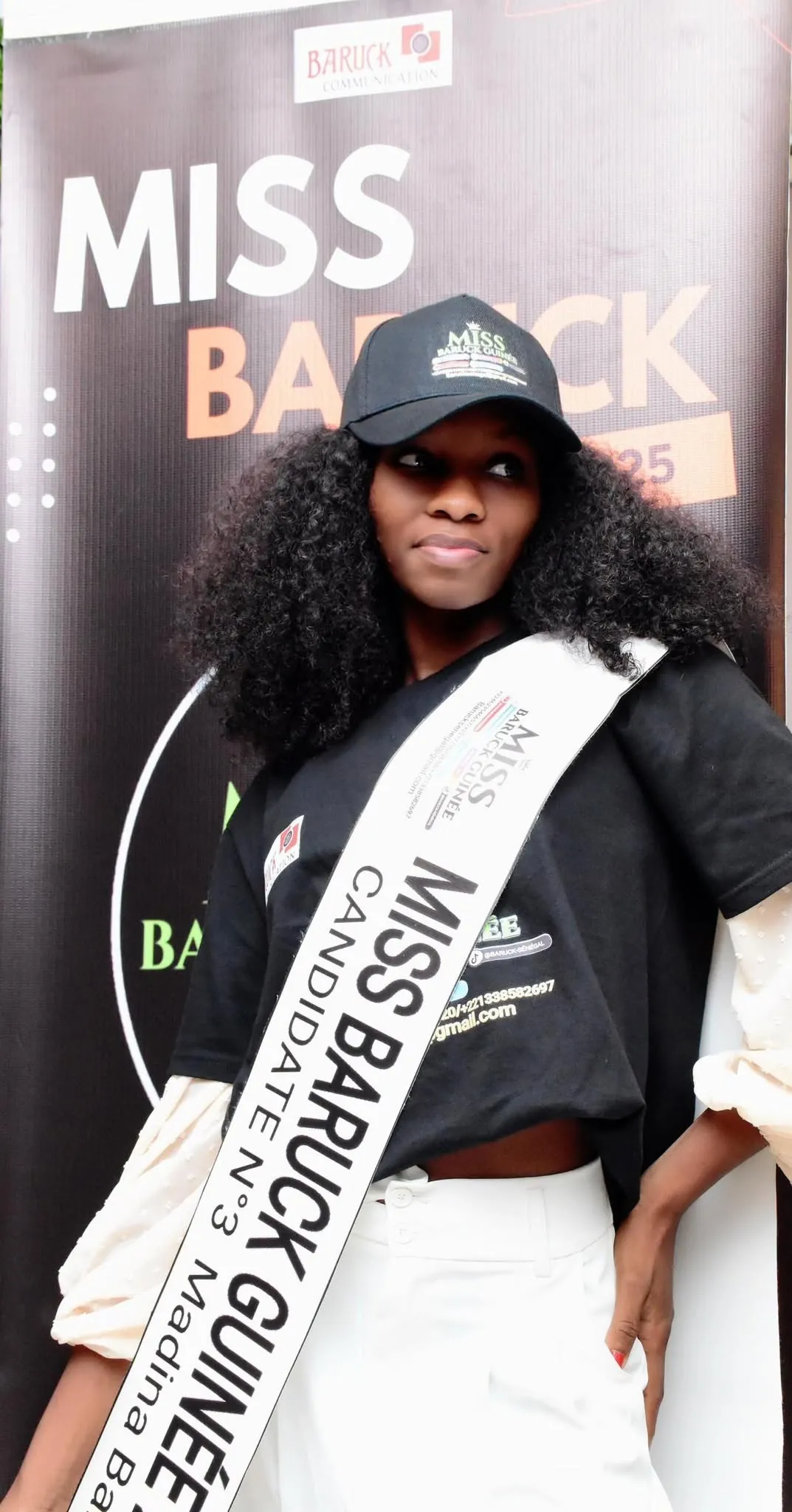 Une candidate en écharpe Miss Baruck Guinée 2025, devant l’affiche de la grande finale
