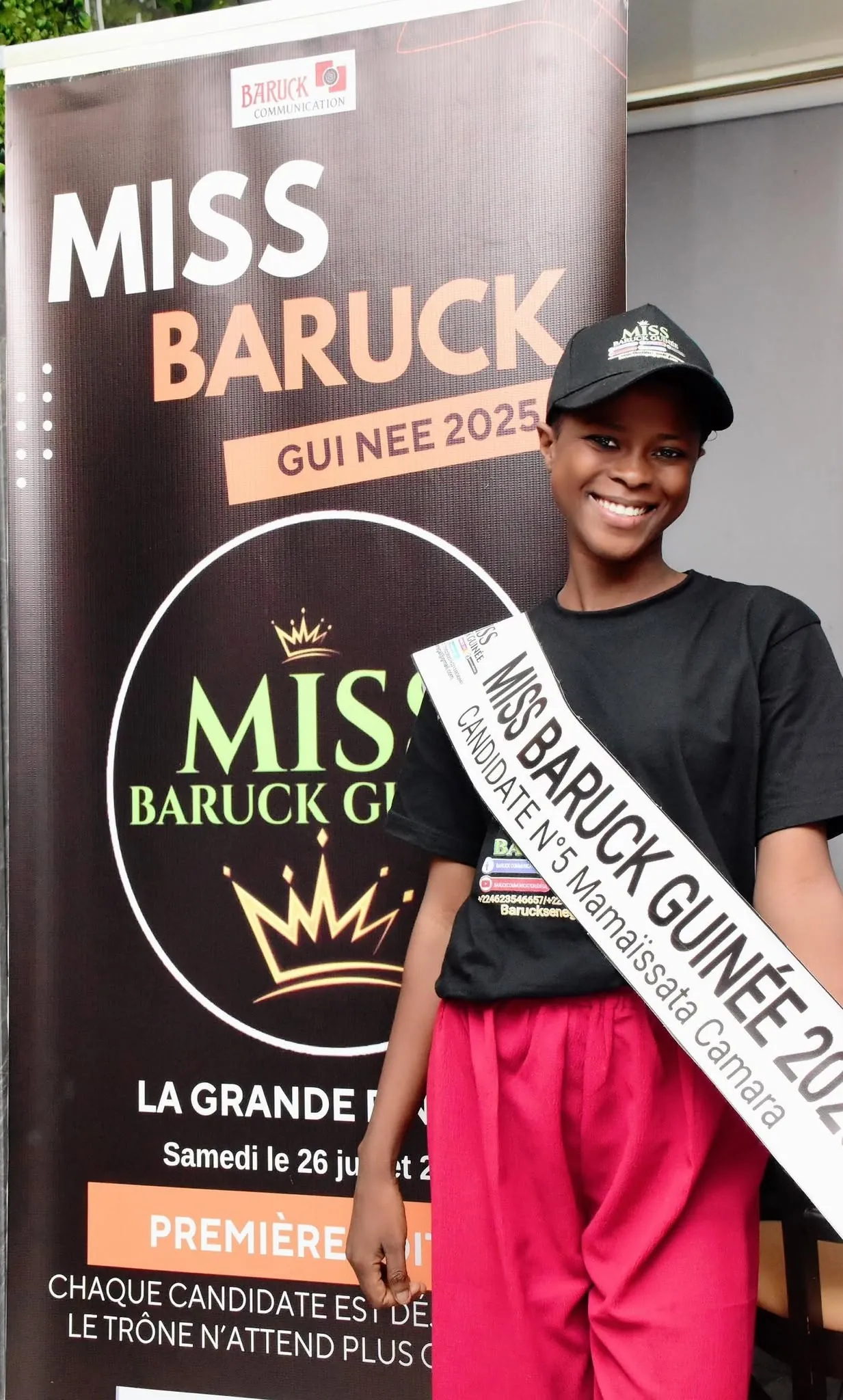 Une candidate souriante en écharpe du concours, devant l’affiche de la première édition
