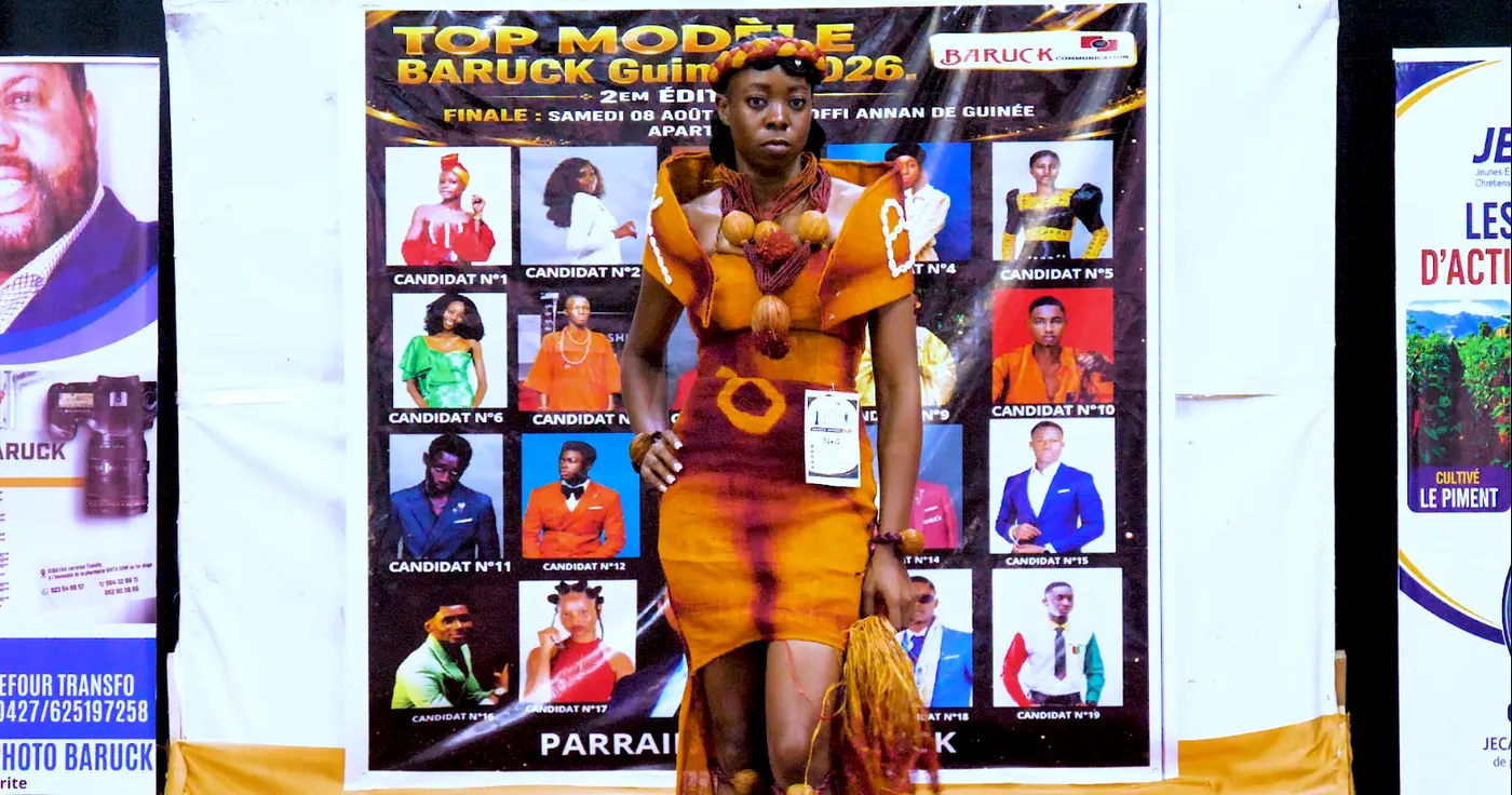 Le candidat n°4 en robe orange brodée, devant l’affiche du concours