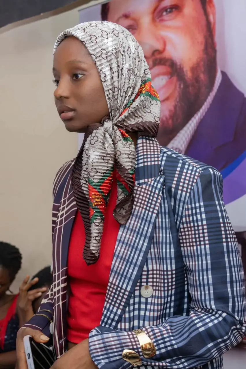 Une candidate en foulard et veste à carreaux, devant l’affiche du PDG du Groupe Baruck